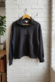 A black quarter-zip pullover sweatshirt hanging on a wooden hanger against a white-painted brick wall in a loft-style room with a window, plant, and rustic wooden furniture