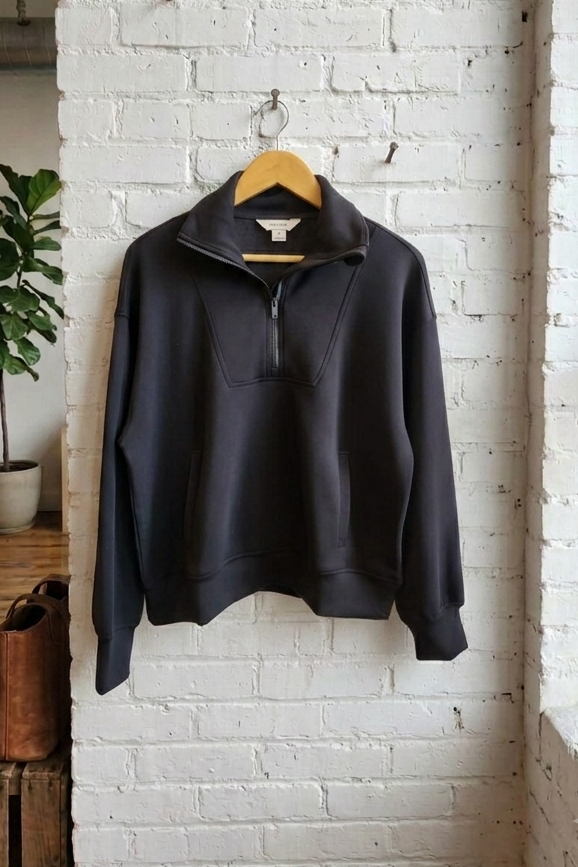 A black quarter-zip pullover sweatshirt hanging on a wooden hanger against a white-painted brick wall in a loft-style room with a window, plant, and rustic wooden furniture