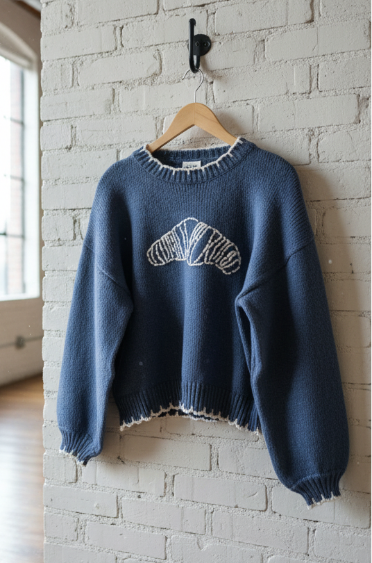 Blue sweater with white croissant logo on a wooden hanger against a white brick wall.