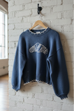 Blue sweater with white croissant logo on a wooden hanger against a white brick wall.