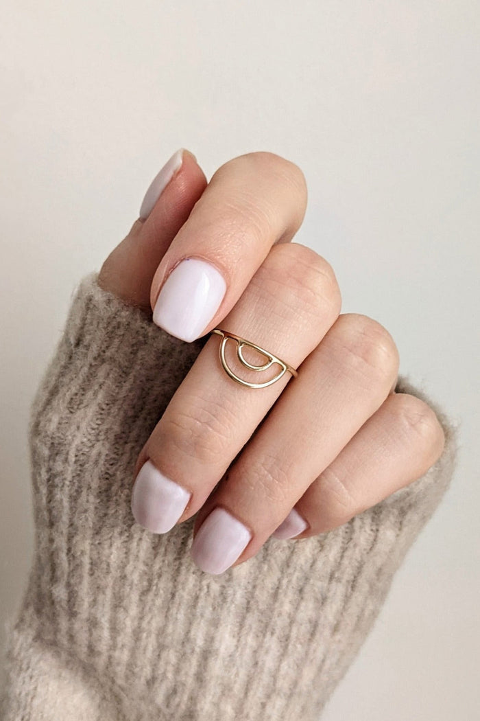 A person's hand wearing a gold-filled arch ring, with a white nail polish and a beige knitted sweater in the background.