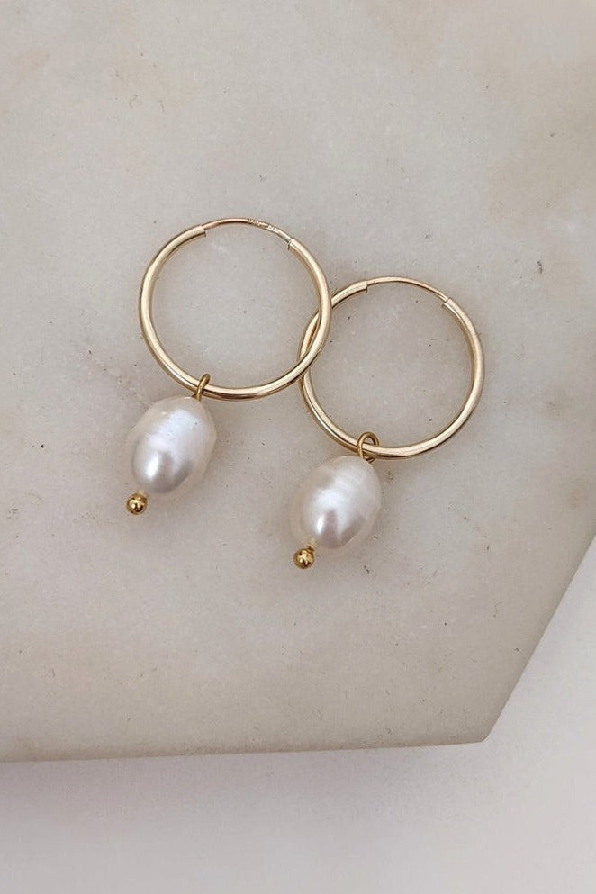 A pair of gold-filled hoop earrings with freshwater pearl drops, displayed on a marble surface.